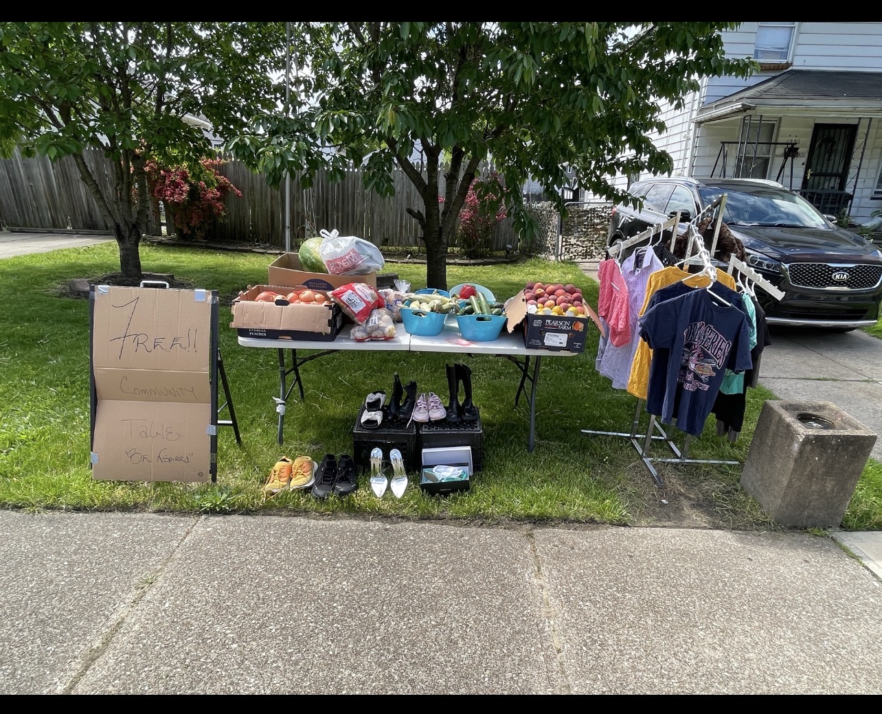 BNspired’s early community table with a handwritten cardboard Free Community Table sign, fresh produce, clothing on a rack, and neatly arranged shoes.